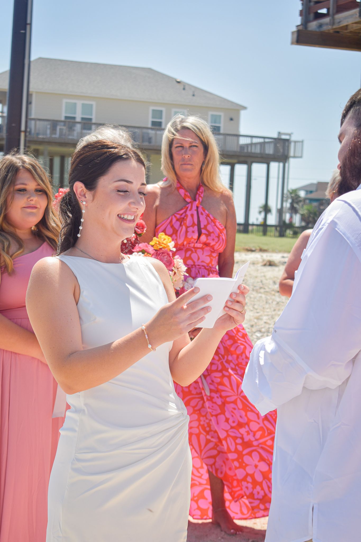 Wedding ceremony on a sunny beach with guests in coral and pink attire celebrating a special moment.