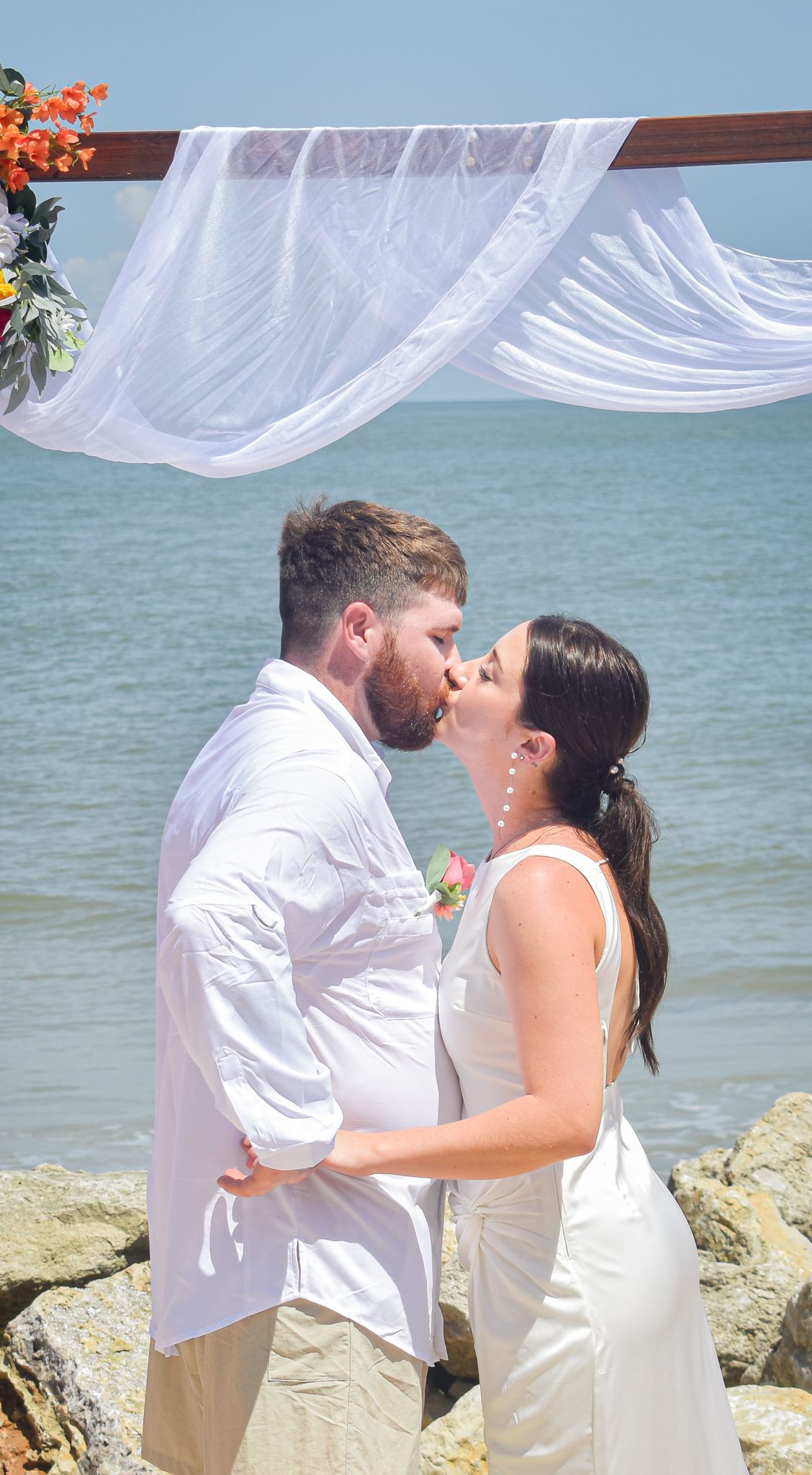 A romantic kiss under a white draped wedding arch on a rocky beach overlooking the ocean.