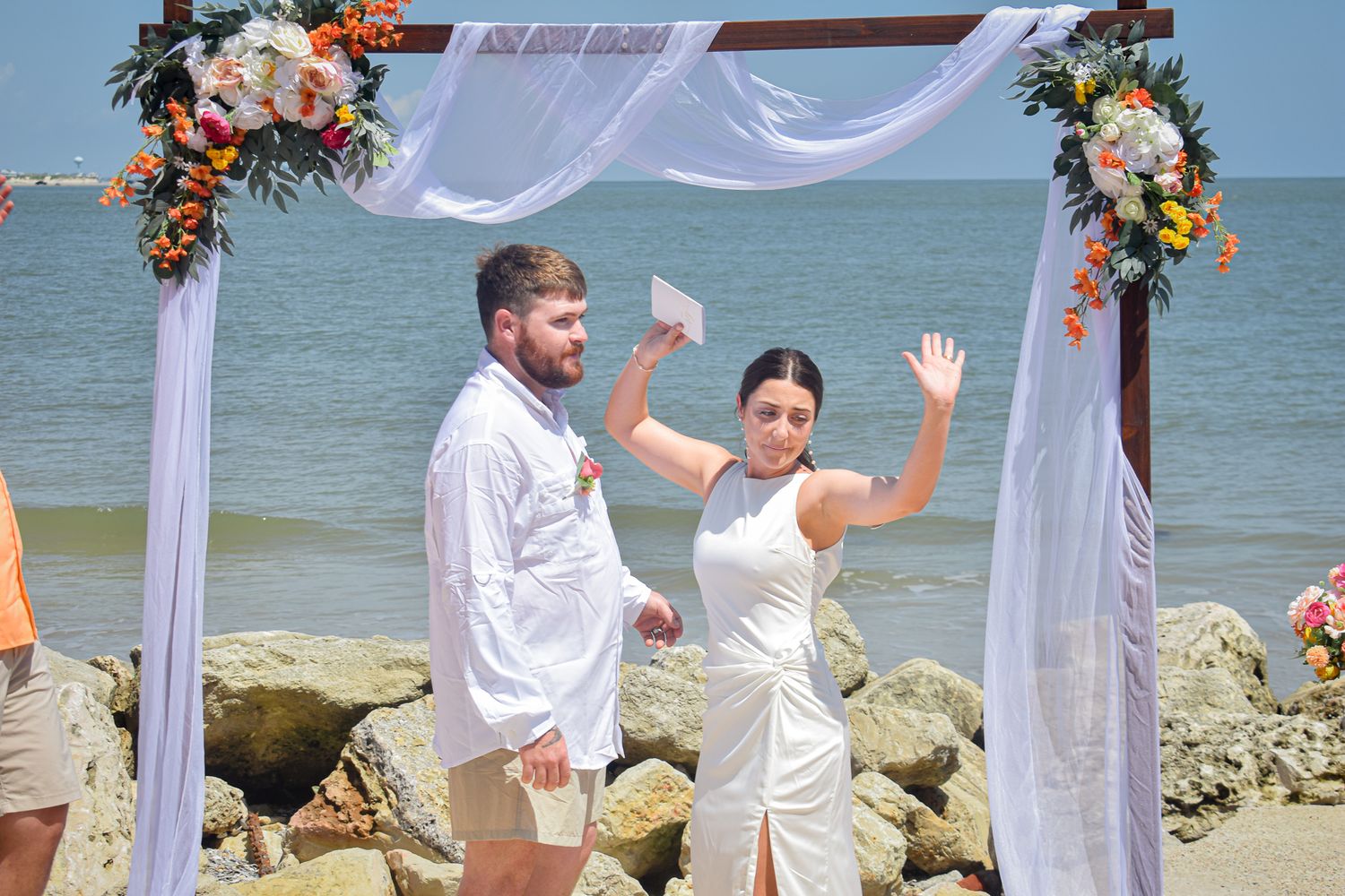 Newlyweds celebrating with joyful gestures under a decorated arch by the seaside.