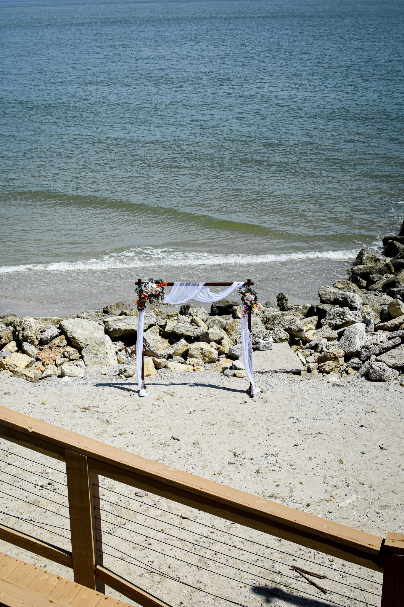 Aerial view of a beach wedding ceremony setup with white arch and rocky shoreline.