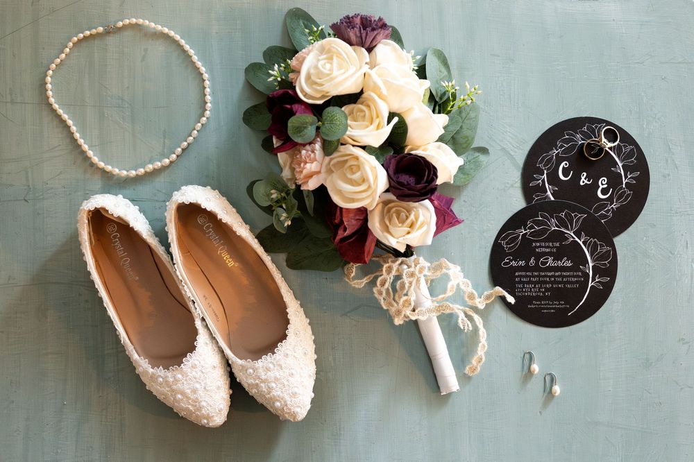 Wedding details flatlay featuring lace flats, pearl necklace, rose bouquet and black invitation cards on sage green surface.