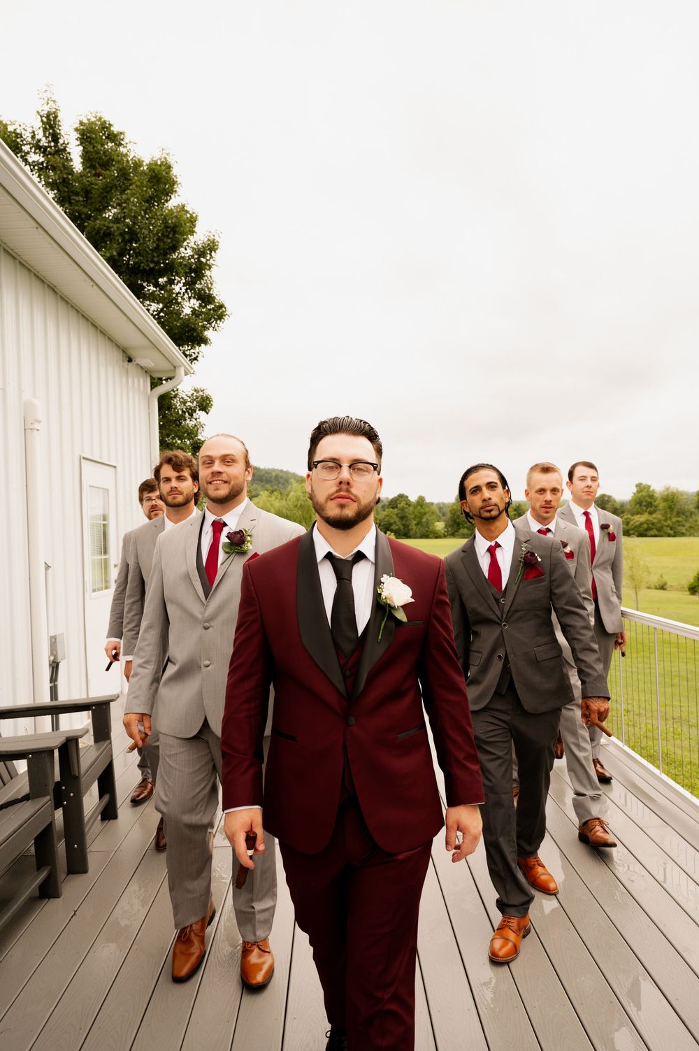 Groomsmen in burgundy and gray suits walk in a line on a white wooden porch with a rural backdrop.