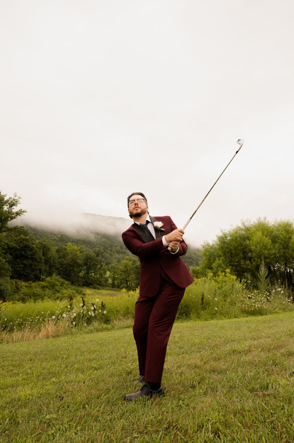 A person in a burgundy suit practices their golf swing on a misty hillside surrounded by green vegetation.
