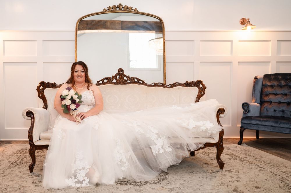 Bride in flowing white wedding gown poses elegantly on antique Victorian sofa beneath ornate mirror in bright room.