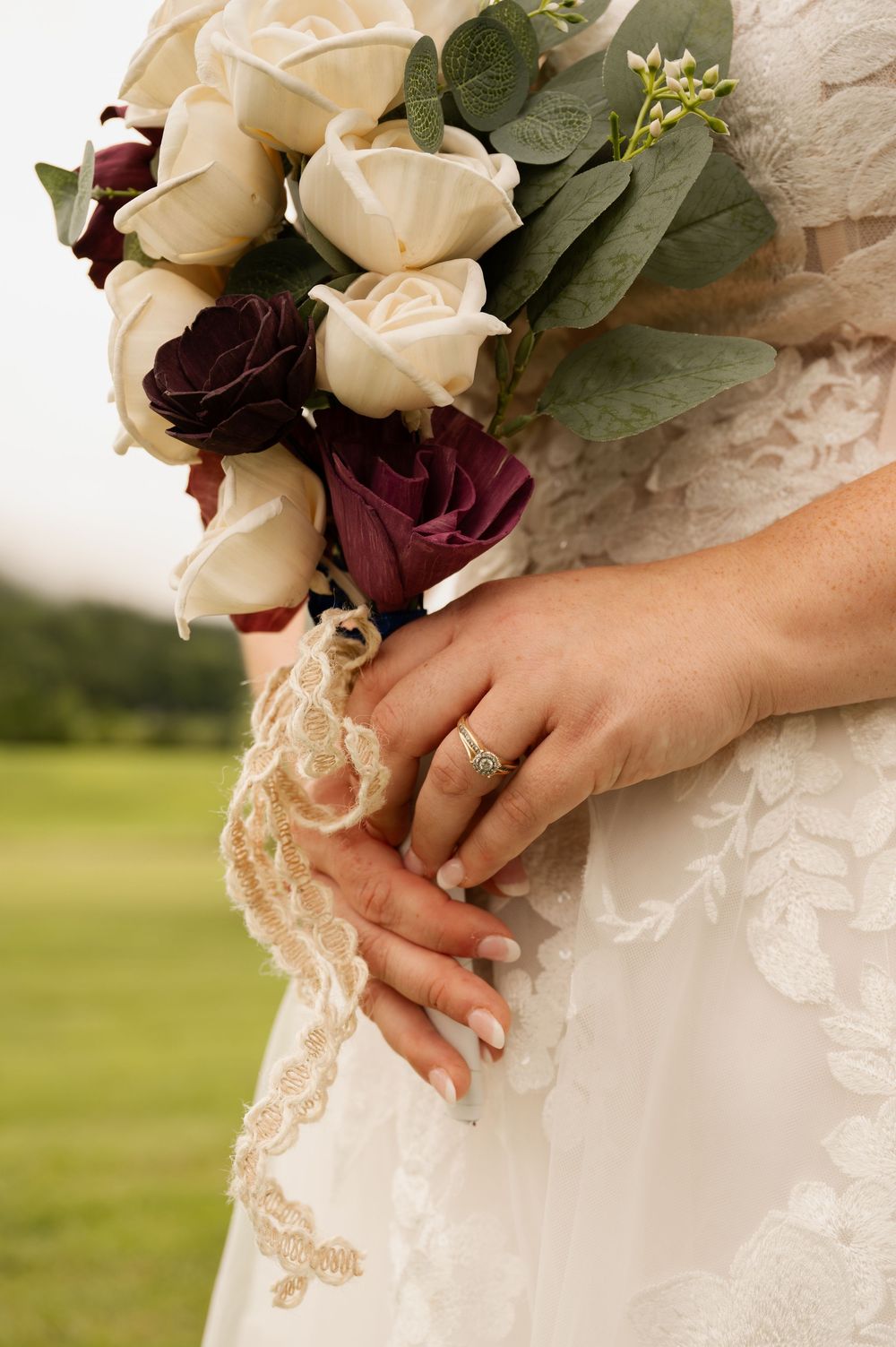 A bridal bouquet of cream and burgundy roses held against a white lace wedding dress with an engagement ring visible.