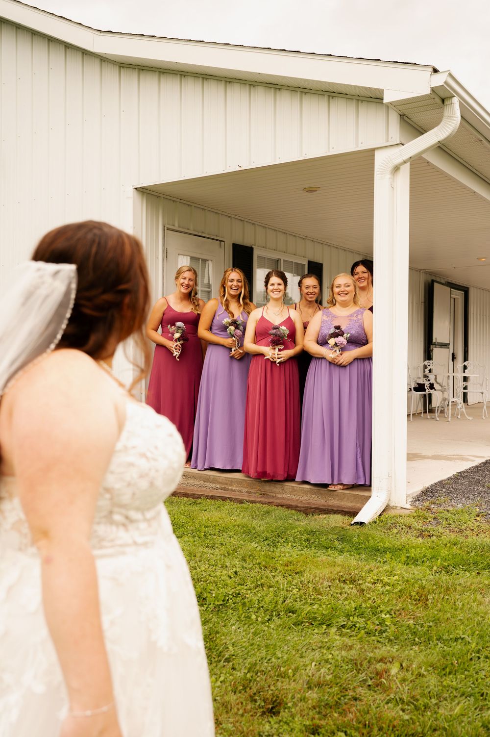 A bride in a white wedding dress stands outside a porch where bridesmaids in purple and burgundy dresses wait.