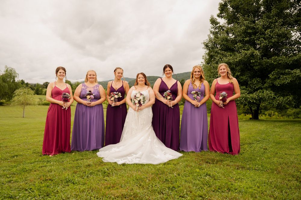 Wedding party in alternating purple and burgundy dresses stand together in a grassy outdoor setting.