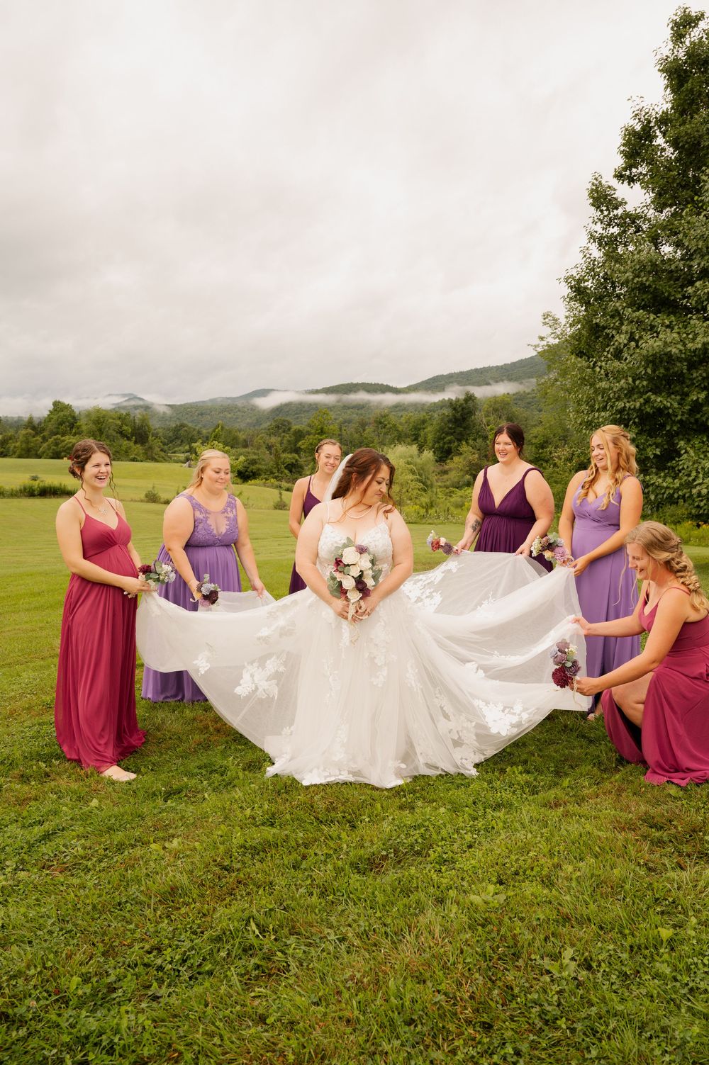 A group in burgundy and purple dresses spread out a white wedding gown on a grassy hillside with mountains in the background.