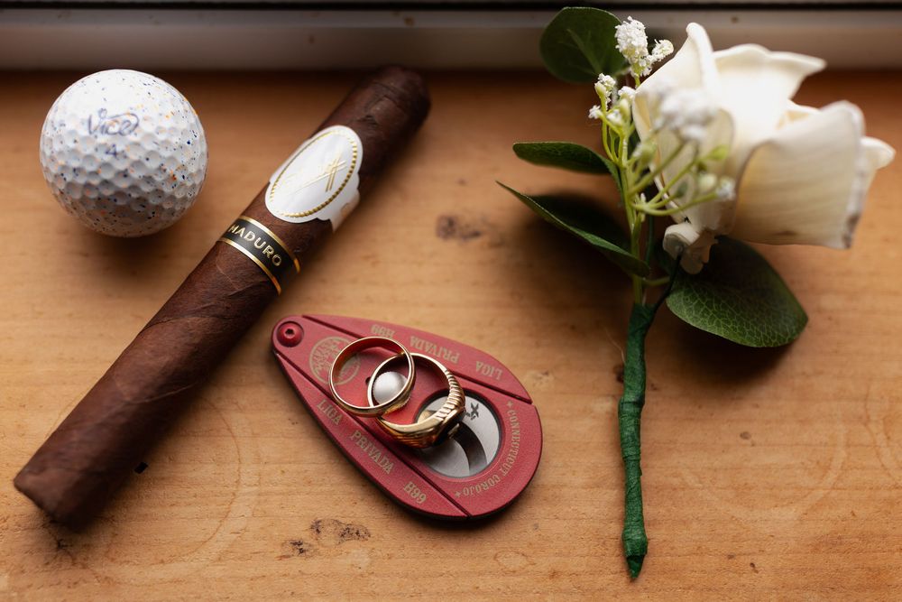 Wedding details including golf ball, cigar, boutonniere, and wedding rings on wooden surface.