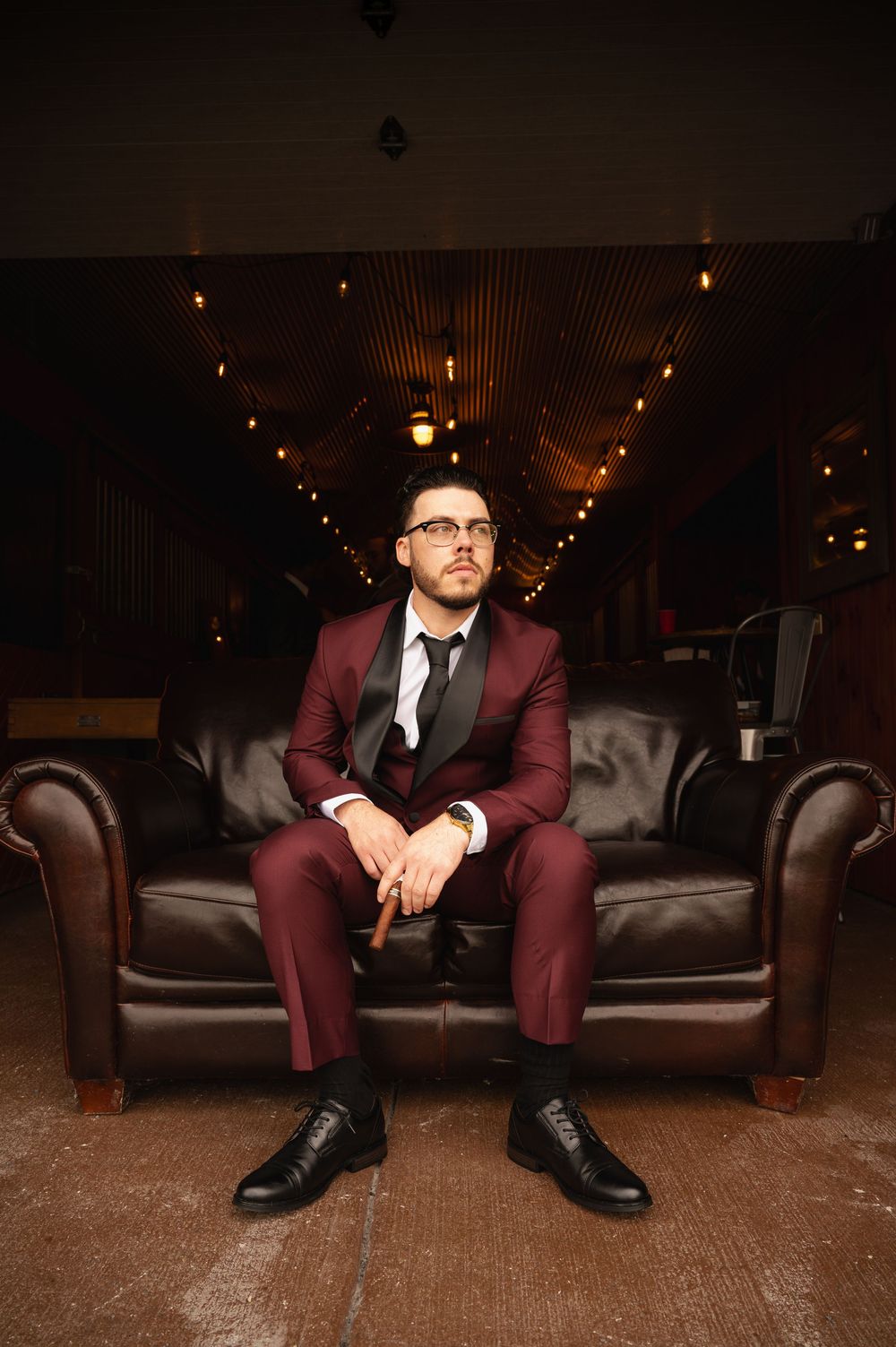 A person in a burgundy suit sits thoughtfully on a dark leather couch under warm string lights in a dimly lit room.