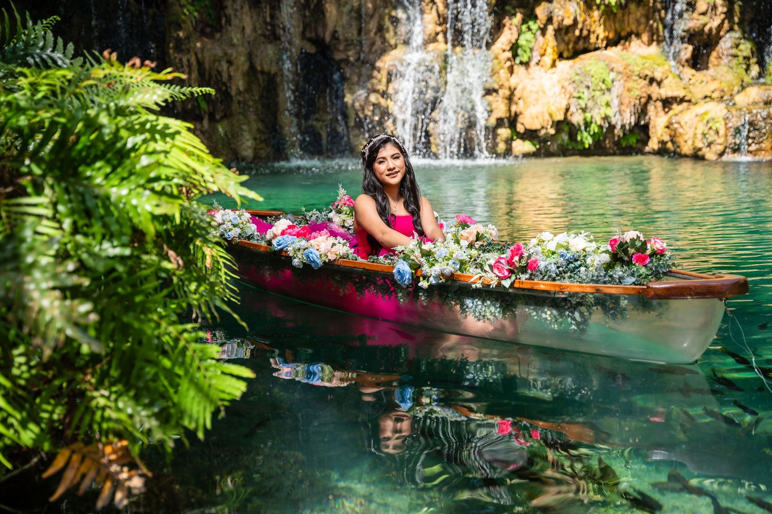 Person in a wooden canoe filled with tropical flowers floats near a scenic waterfall and turquoise lagoon surrounded by ferns.