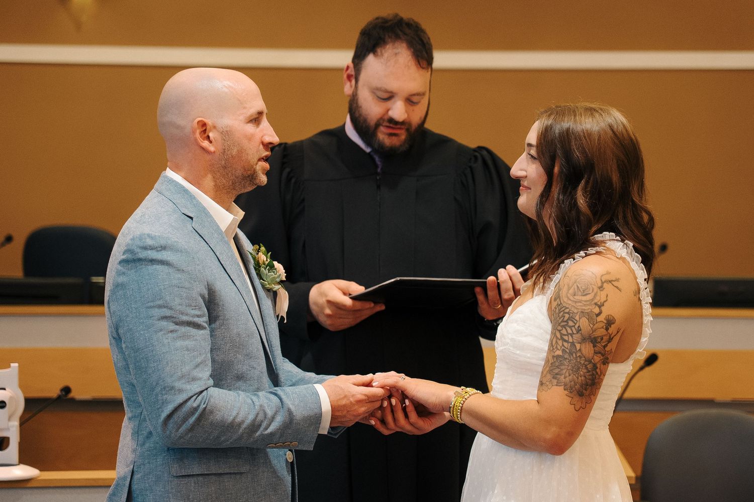 An intimate wedding ceremony takes place with the couple exchanging rings as an officiant in black robes presides.