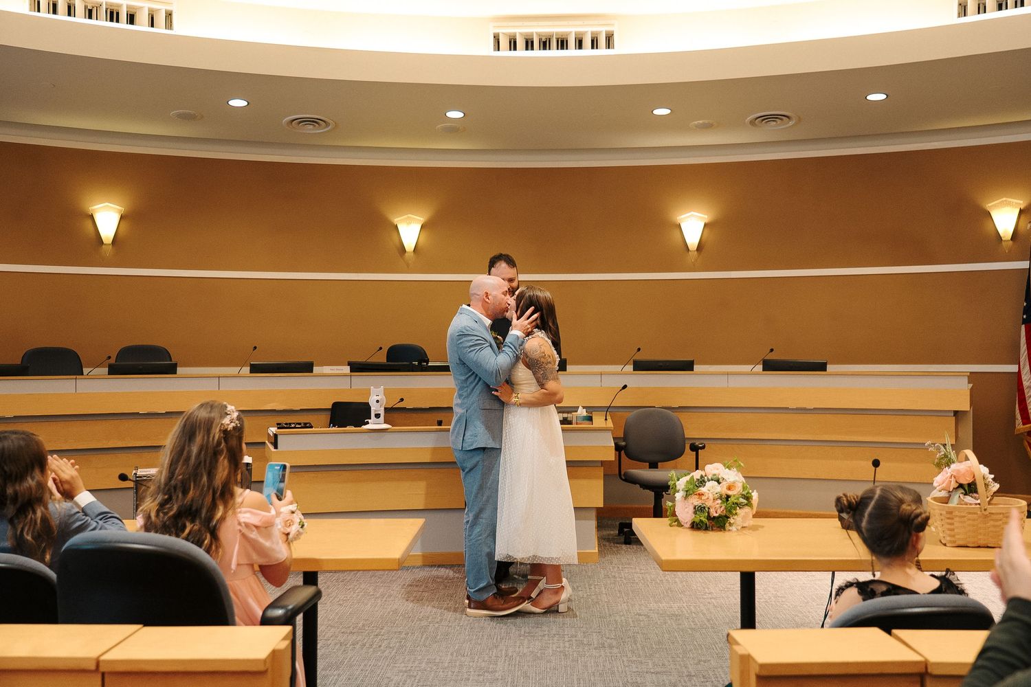 A couple shares their first dance in a courthouse ceremony room while guests look on from wooden benches.