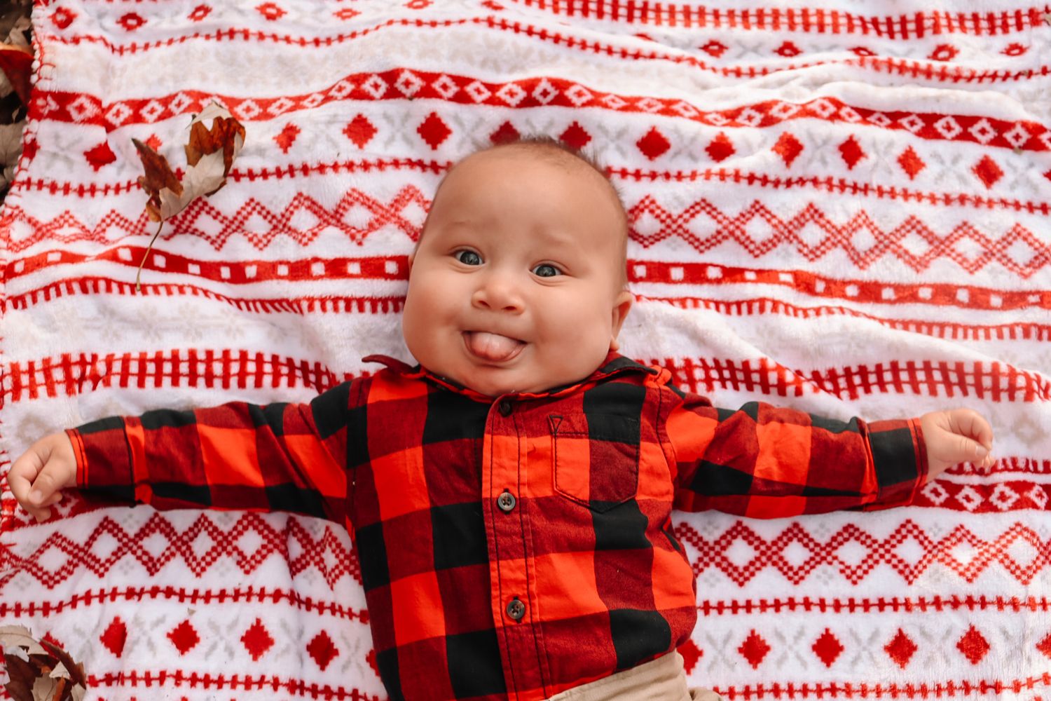 A small child wearing a red buffalo plaid shirt lies on a white and red patterned blanket.