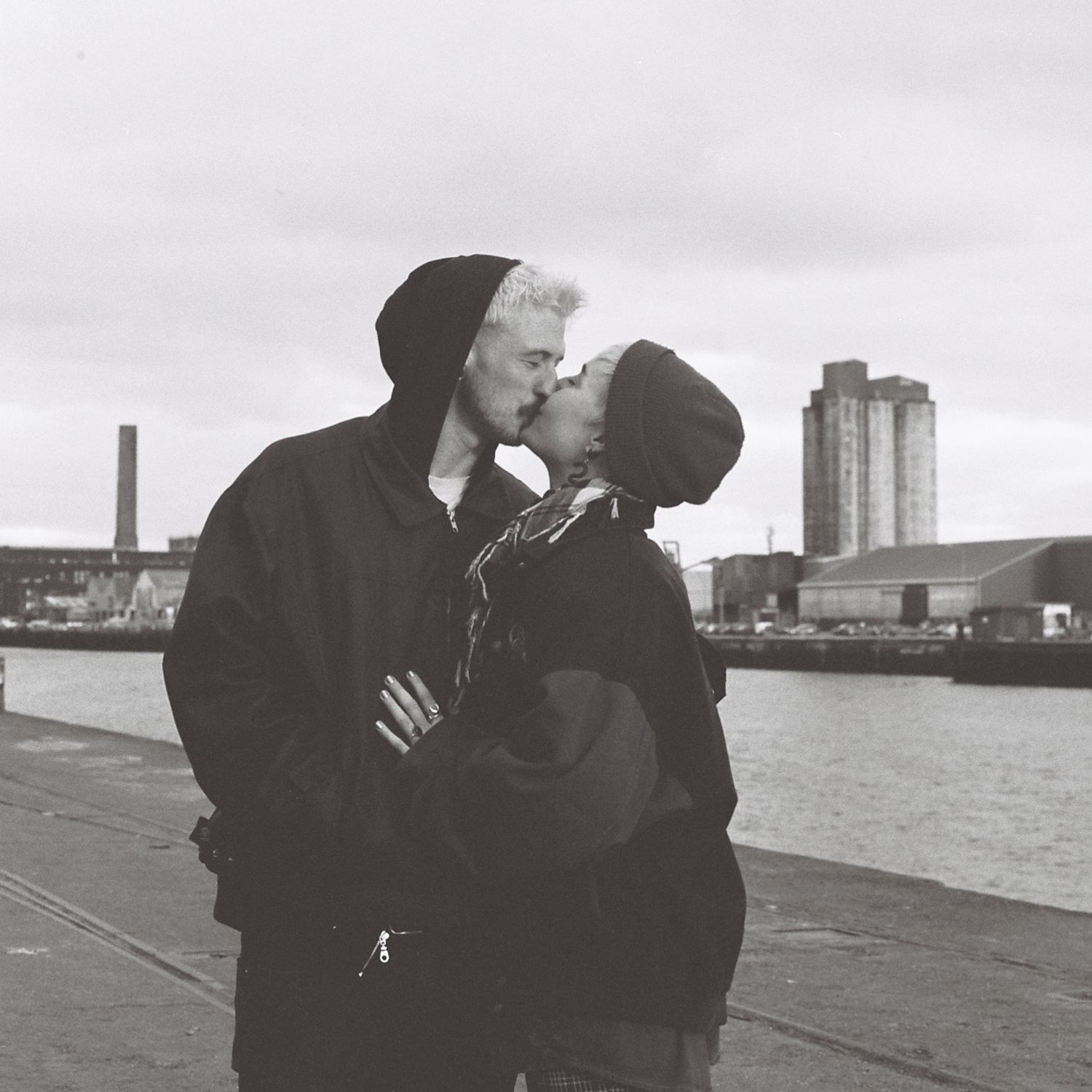 Two people share an intimate moment by the waterfront on an overcast day.