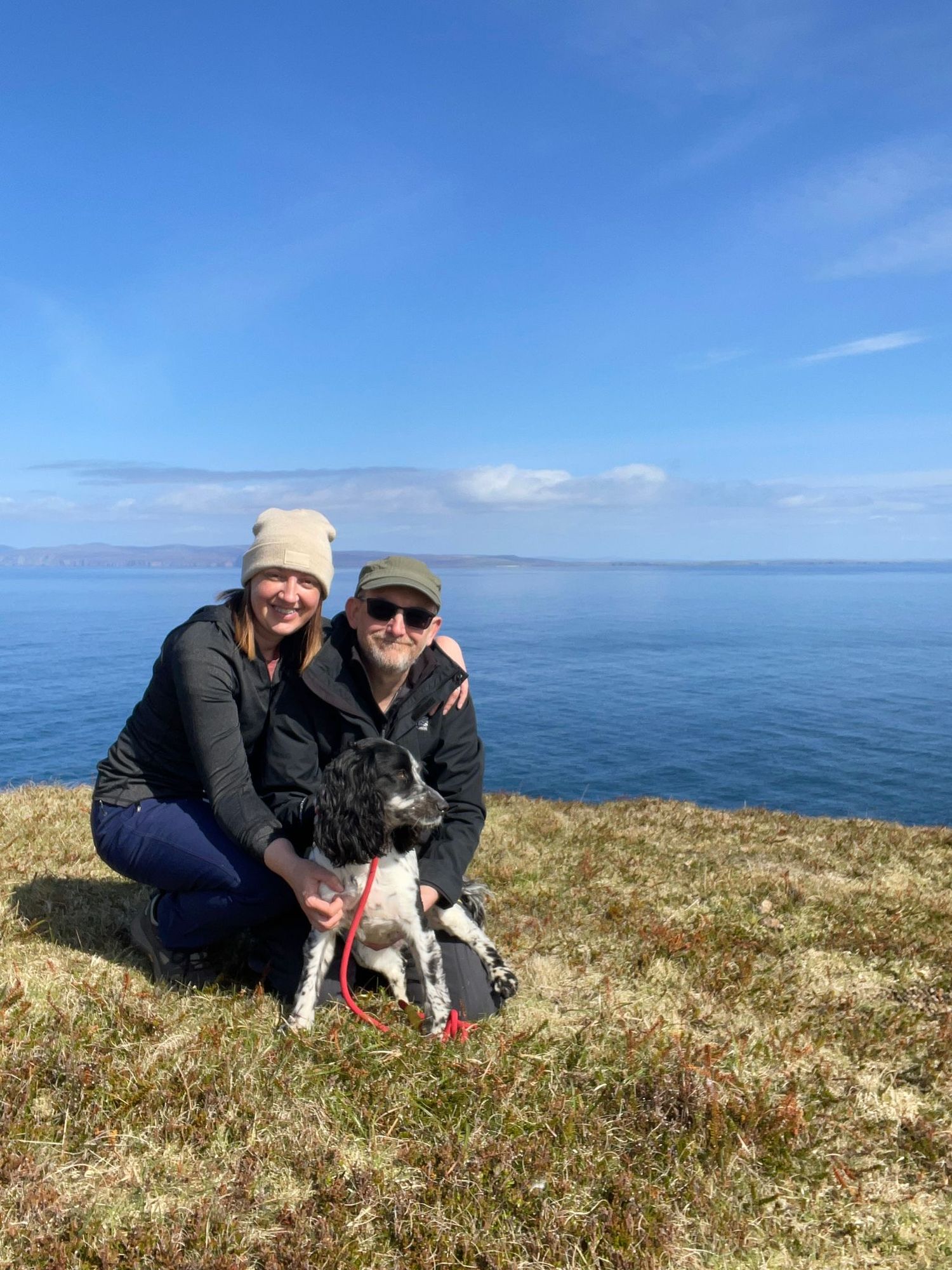 a couple and their cocker spaniel sat on the cliff top of dune head in the highlands of Scotland. they are crouched down fussing their dog.