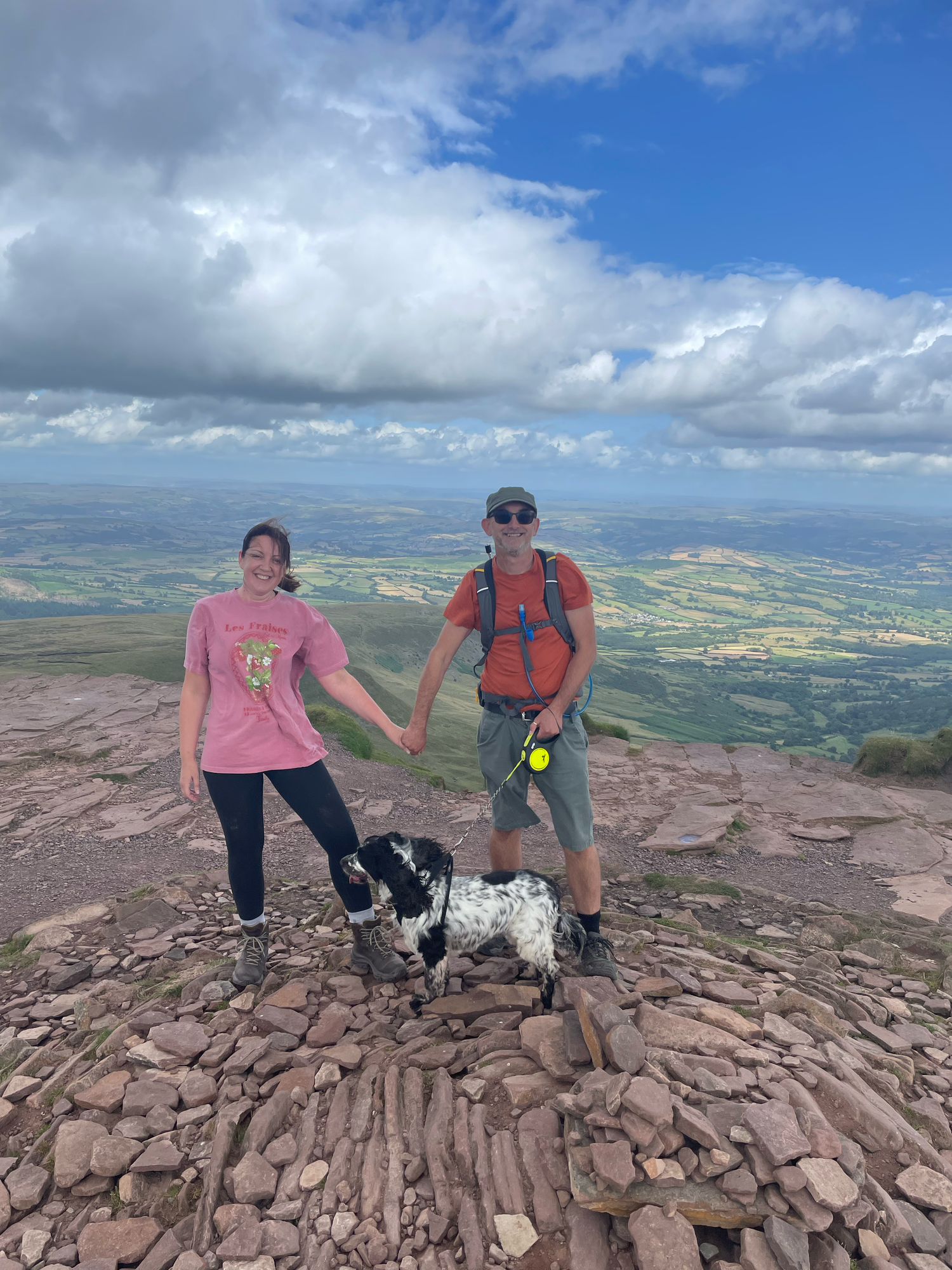 a couple are stood at the top of Corn Dun mountain trig point. they are holding hands and a view of the welsh valley is in the background