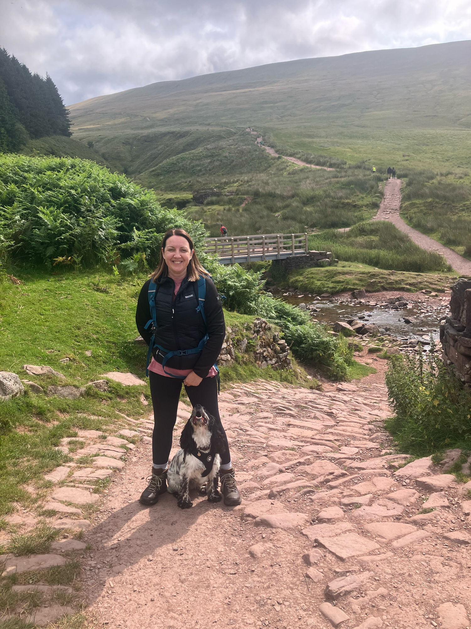 a lady in hiking clothing is stood at the Botton of Pen-Y-Fan mountain with her cocker spaniel.