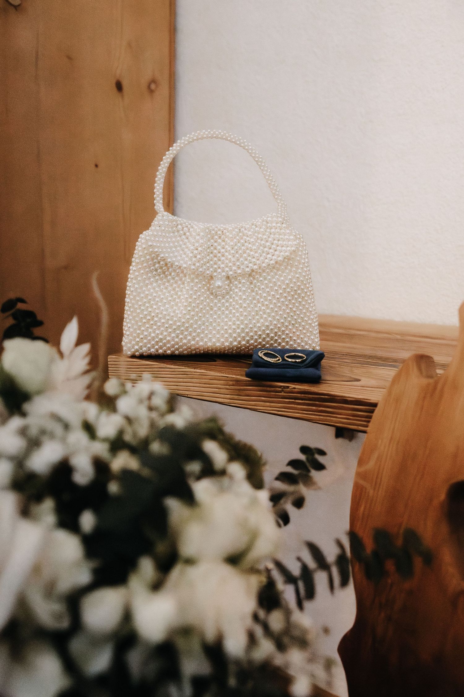Vintage white beaded handbag sits on wooden shelf next to delicate white flowers and eucalyptus.