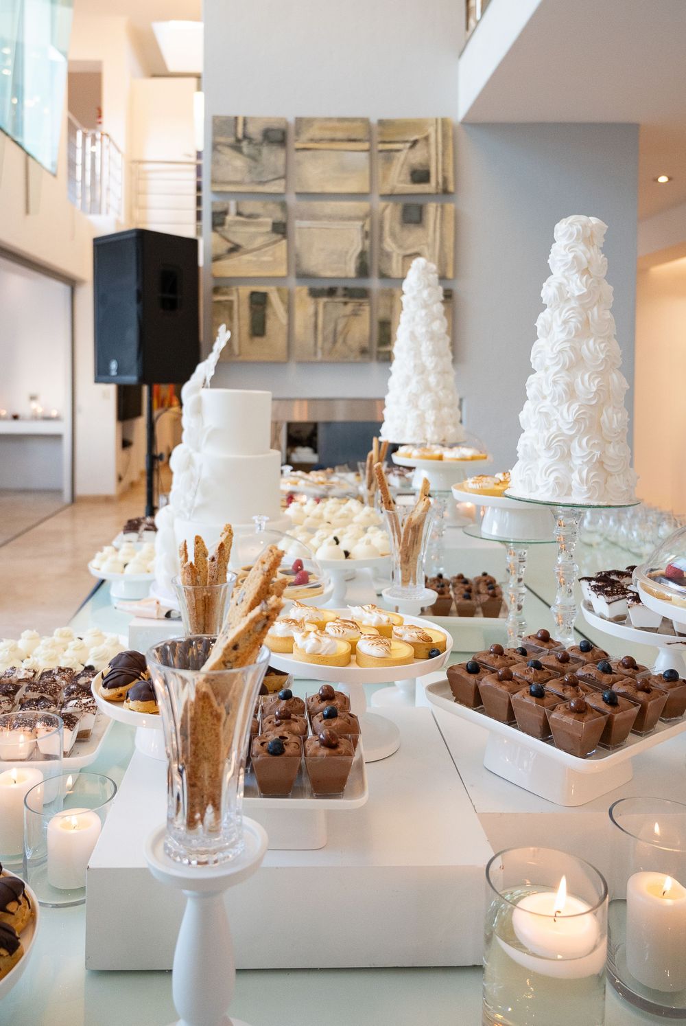 Luxurious dessert table display featuring tall white wedding cakes, chocolate treats, and glowing candles on white platforms.