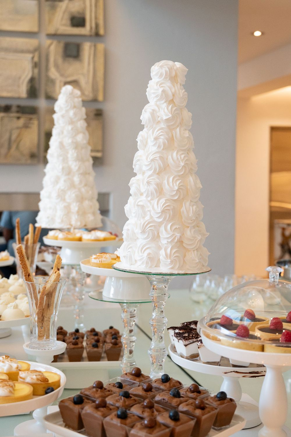 Elegant white meringue tower cakes and chocolate desserts displayed on glass stands at a luxury dessert buffet table.