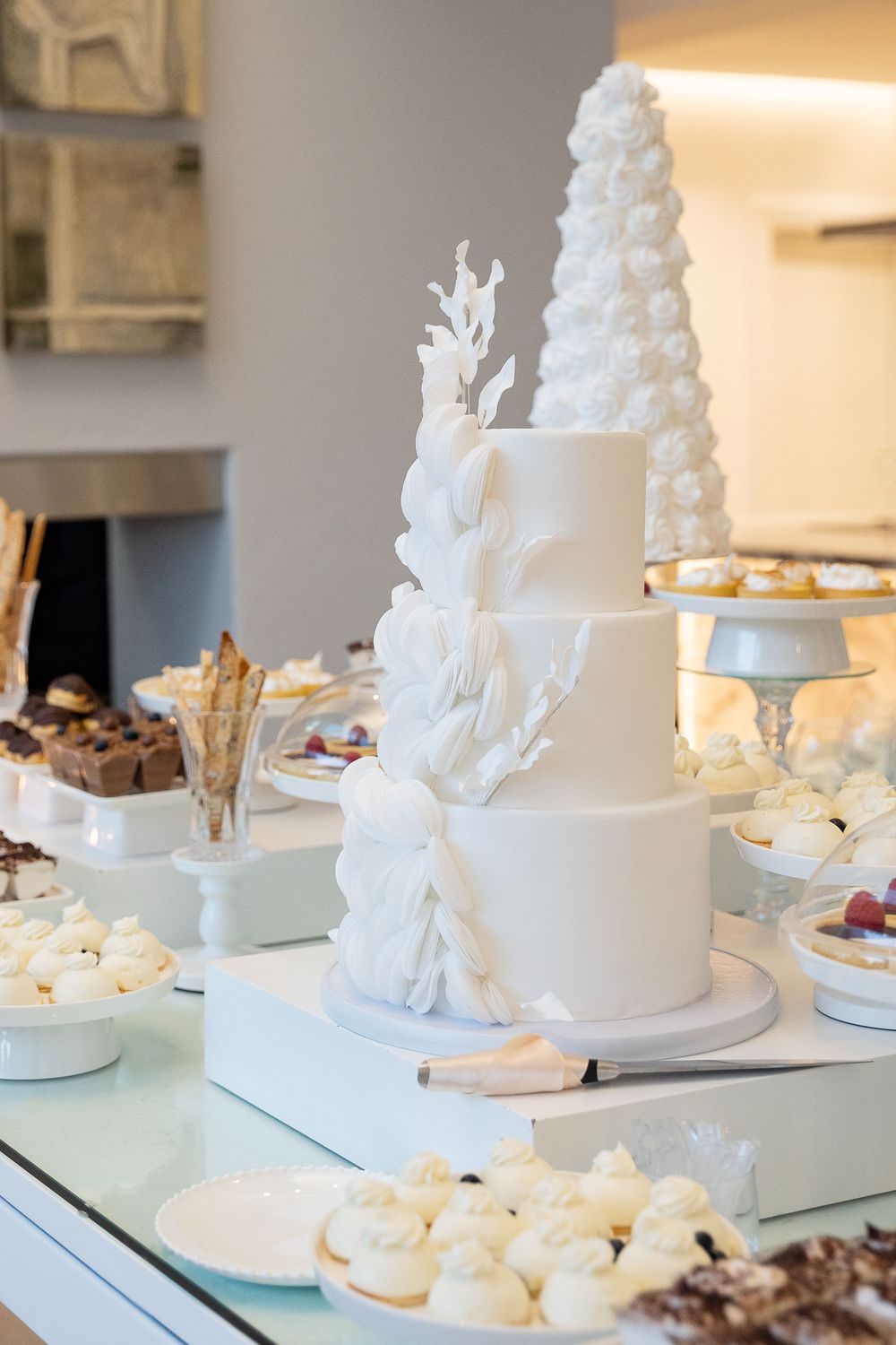 Elegant white wedding cake decorated with coral-inspired details sits on dessert table with assorted sweet treats.