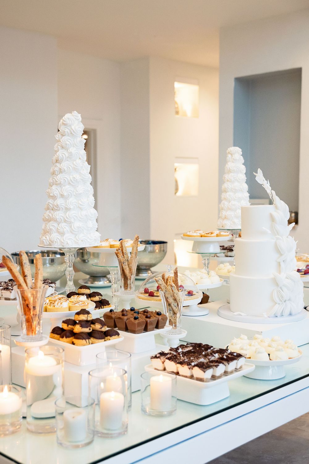 Elegant wedding dessert table display with tall white tiered cakes and assorted treats on glass shelving with candlelight.