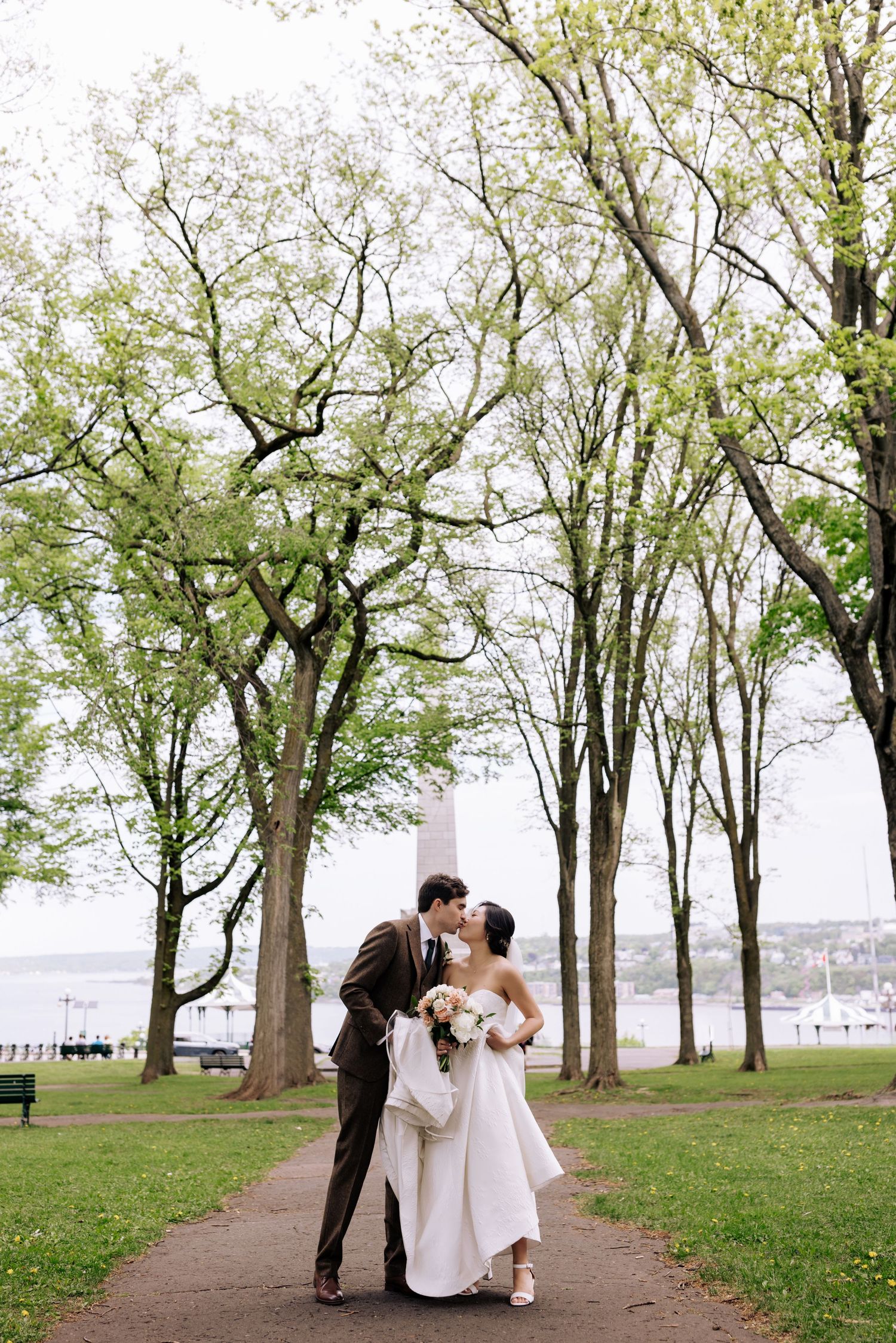 A couple shares an intimate moment on a tree-lined path near a waterfront.