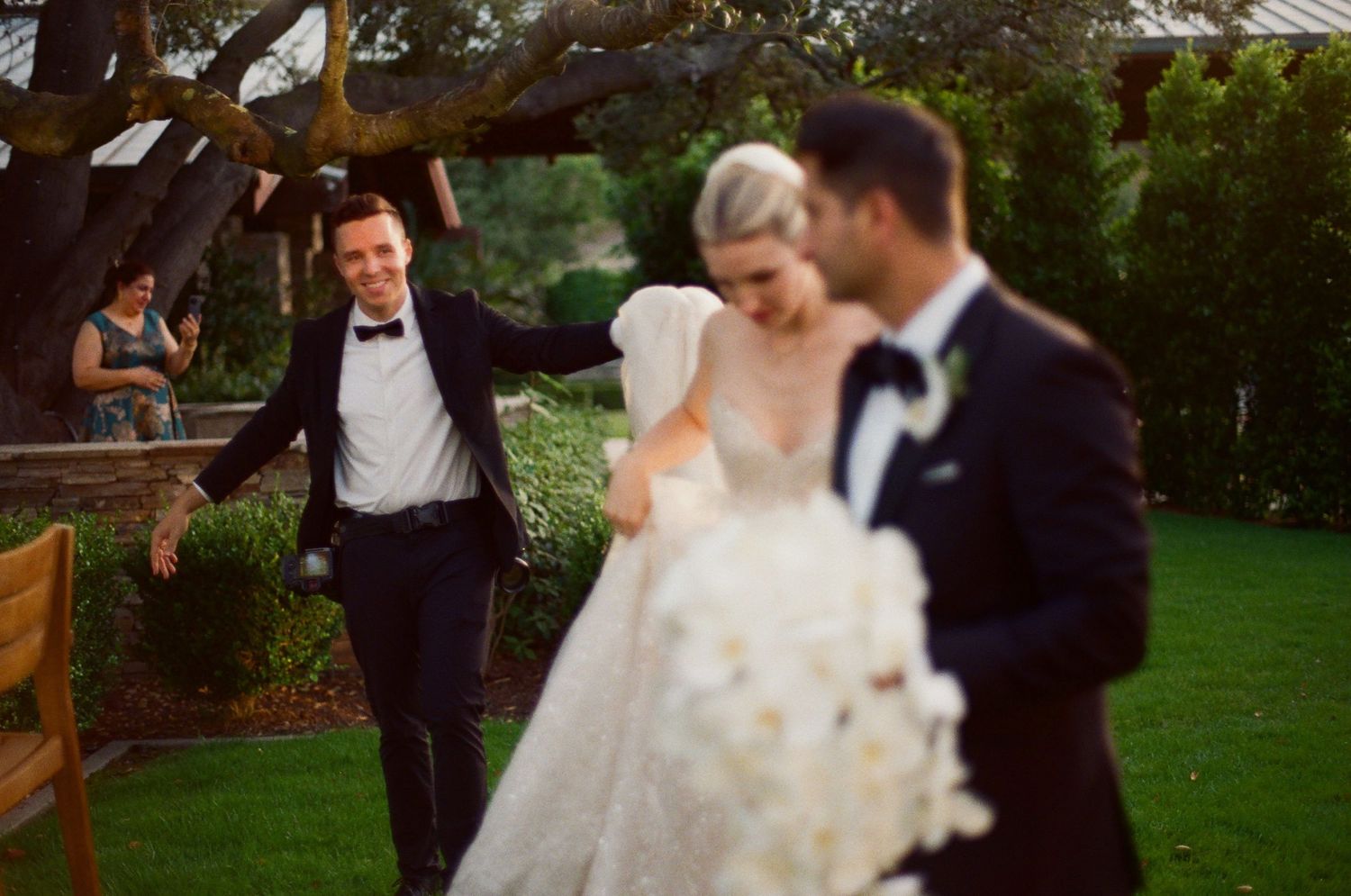 A cheerful groomsman makes a playful entrance behind a couple during an outdoor garden wedding ceremony.