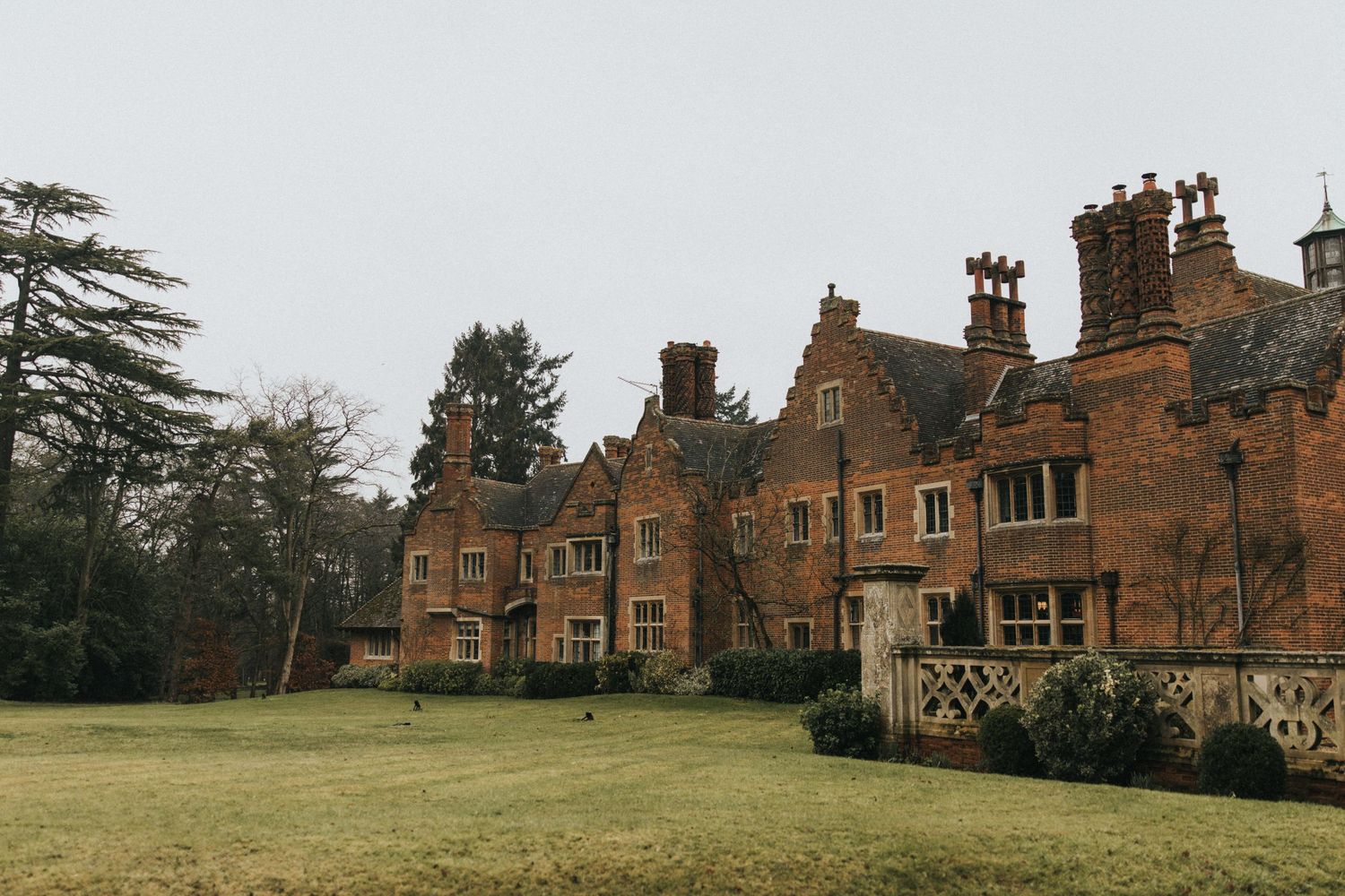 Historic English manor house with red brick exterior and multiple chimneys sits beside a sprawling green lawn.
