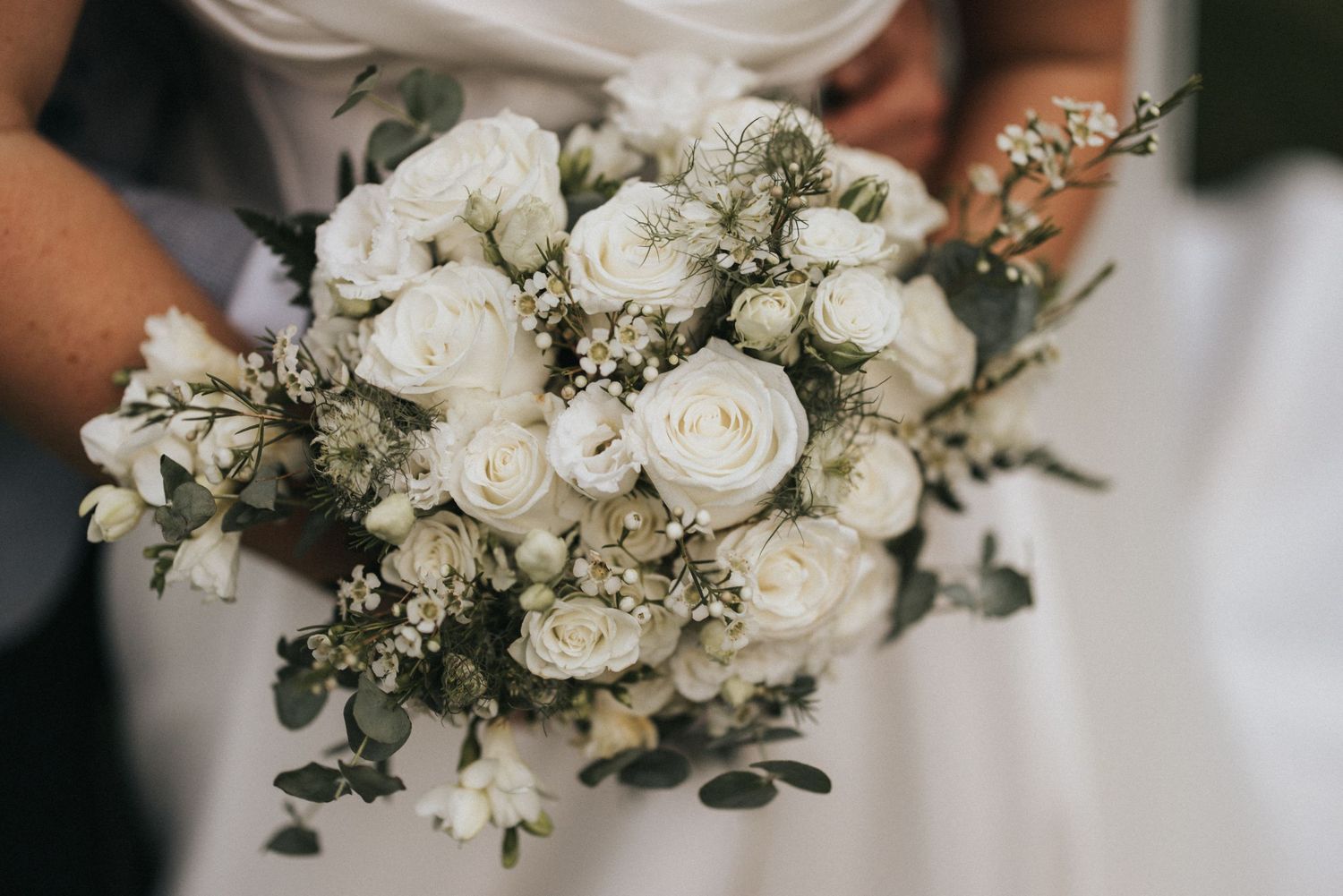 A stunning white wedding bouquet featuring roses, eucalyptus leaves and baby's breath held against a white dress.