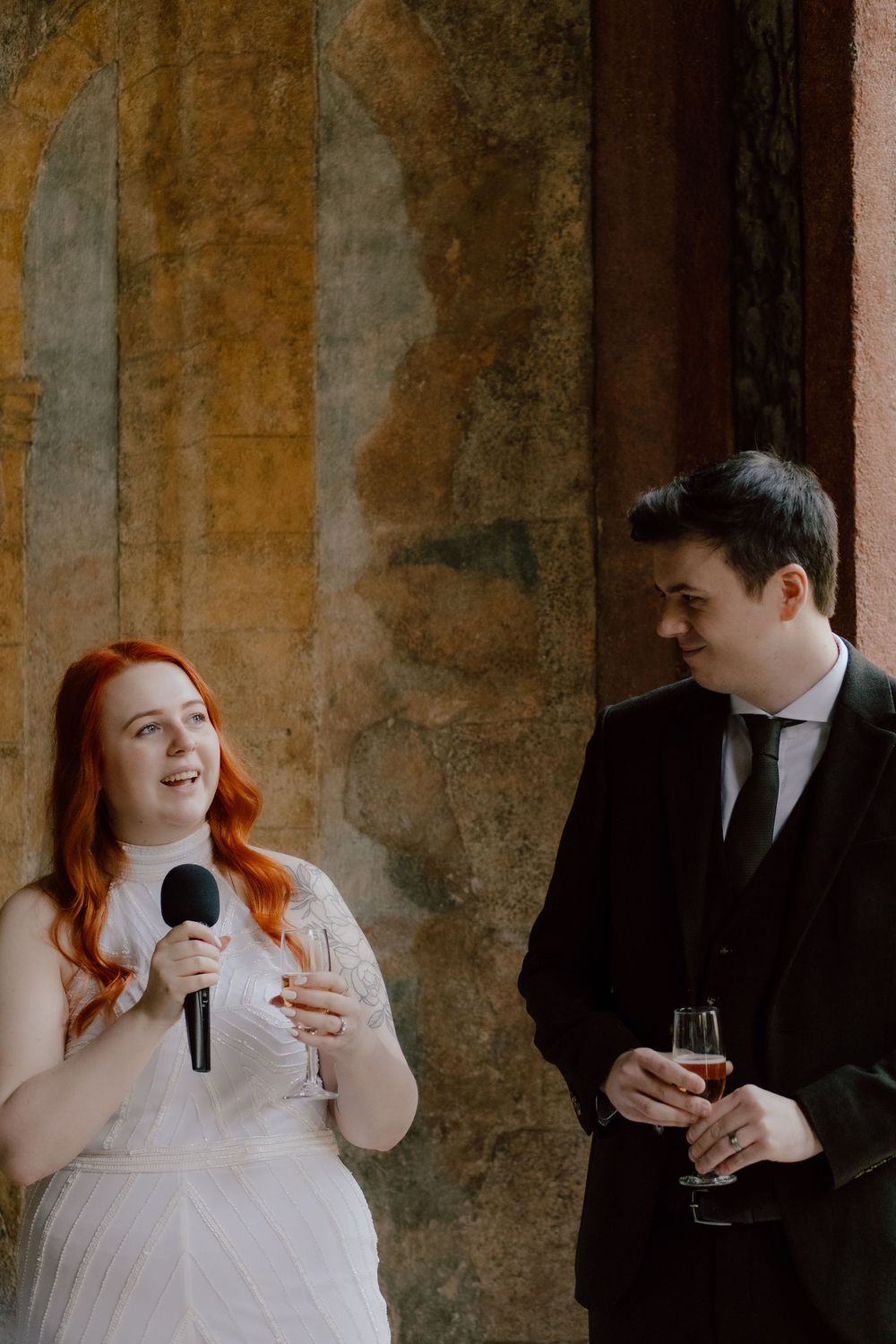 Joyful wedding couple sharing intimate moment and laughter while holding glasses in elegant rustic venue.