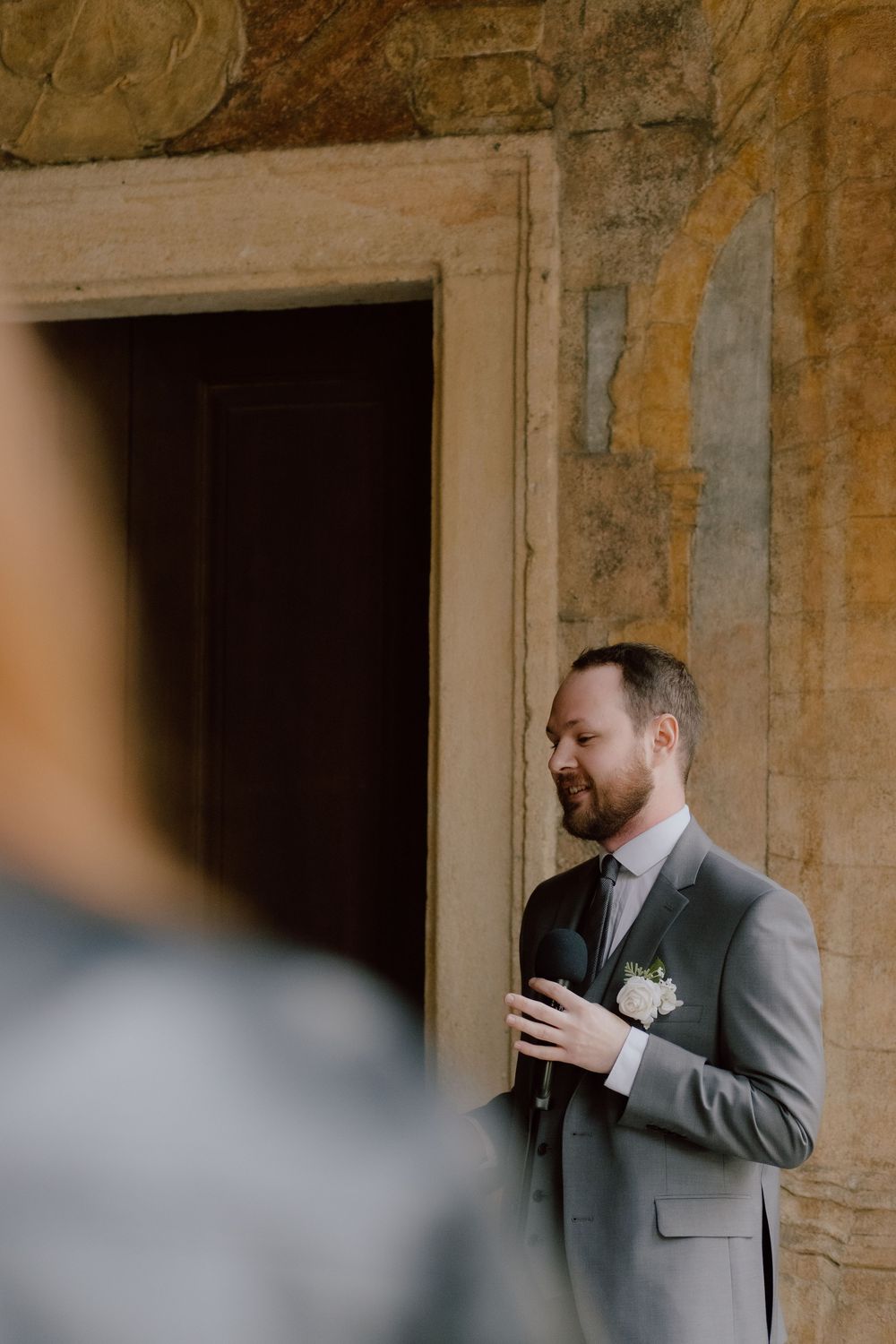 A groom in a grey suit stands in an elegant stone doorway during a wedding ceremony.