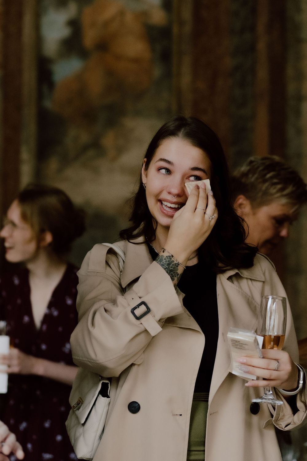 A guest in a beige trench coat laughs while holding a champagne glass at an outdoor event.