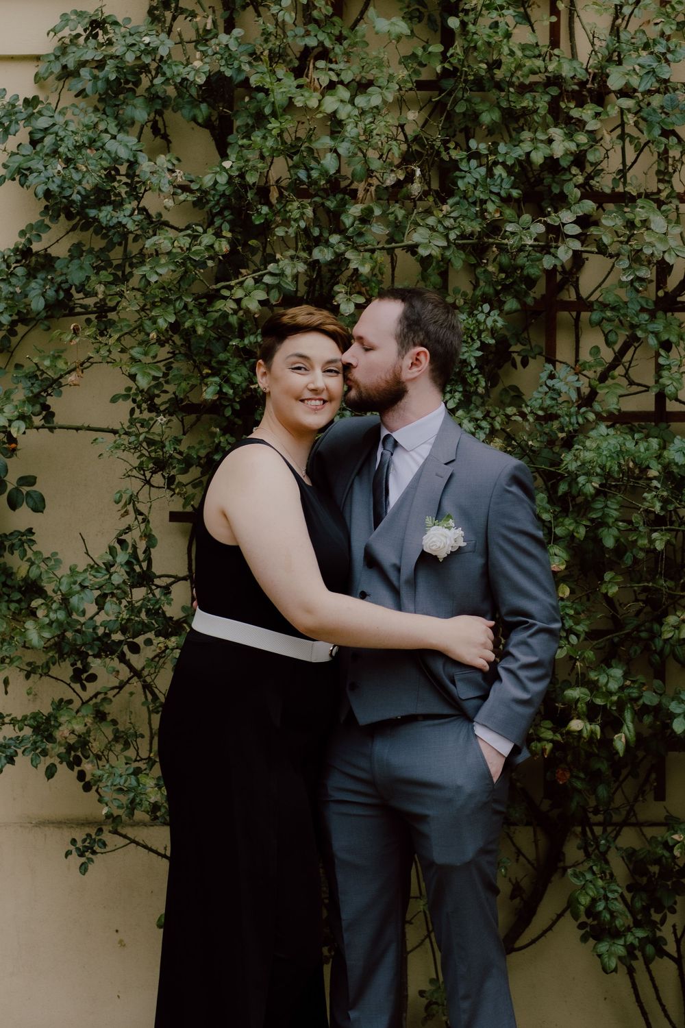 A romantic couple embraces against a wall covered in lush green vines during a wedding celebration.