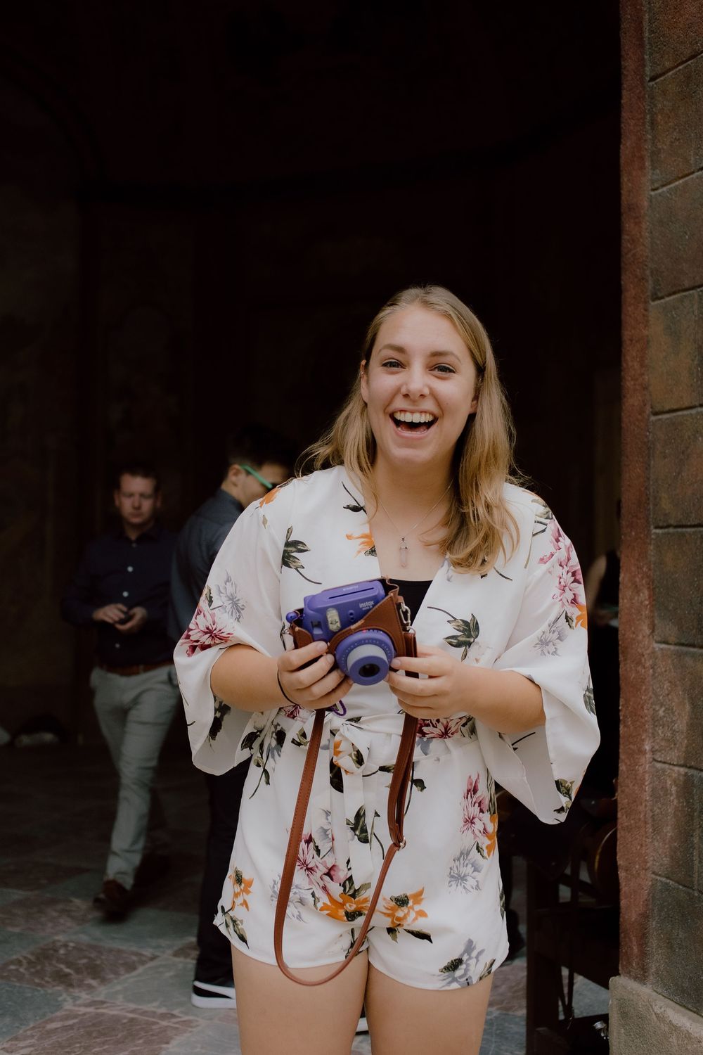 Person in floral print dress laughing while holding a camera in stone archway setting.