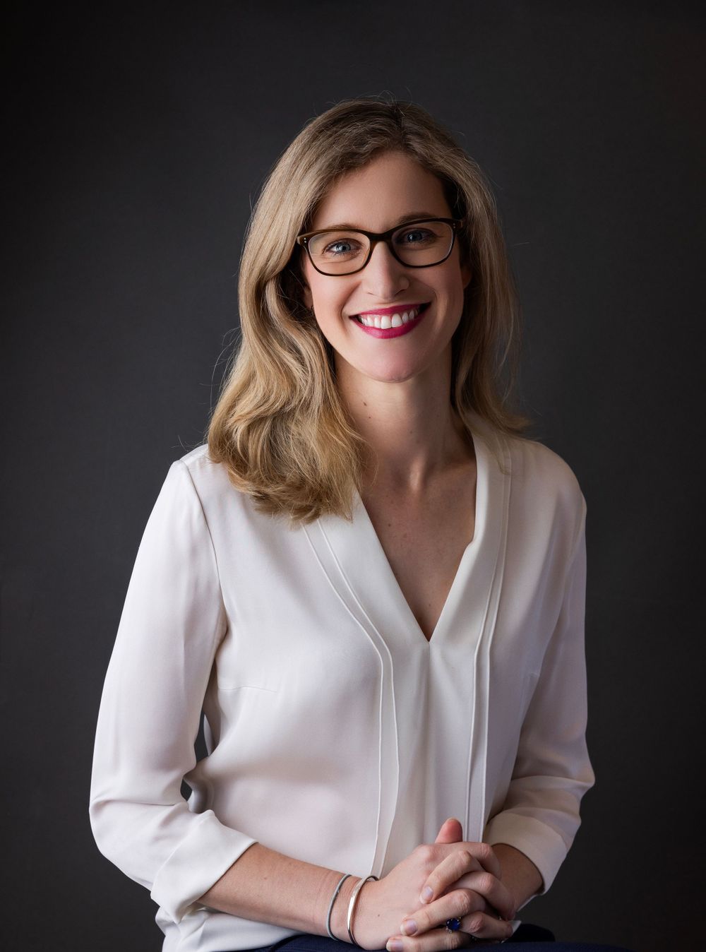 Professional woman in white blouse and glasses smiling against dark backdrop.