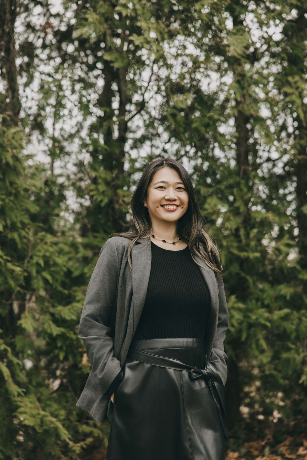 A person wearing a gray blazer and black top poses outdoors against a backdrop of green trees in natural lighting.