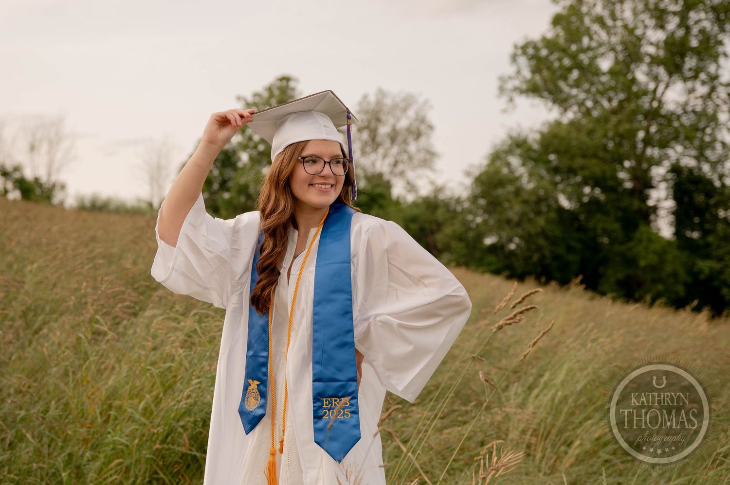 Graduate wearing white cap and gown with blue stole poses joyfully in a grassy meadow during golden hour.