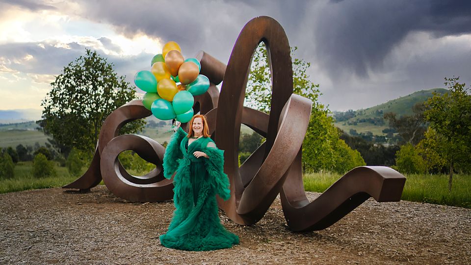 Large sculpture installation in a park captures a flowing metal ribbon form against dramatic cloudy skies and mountains.