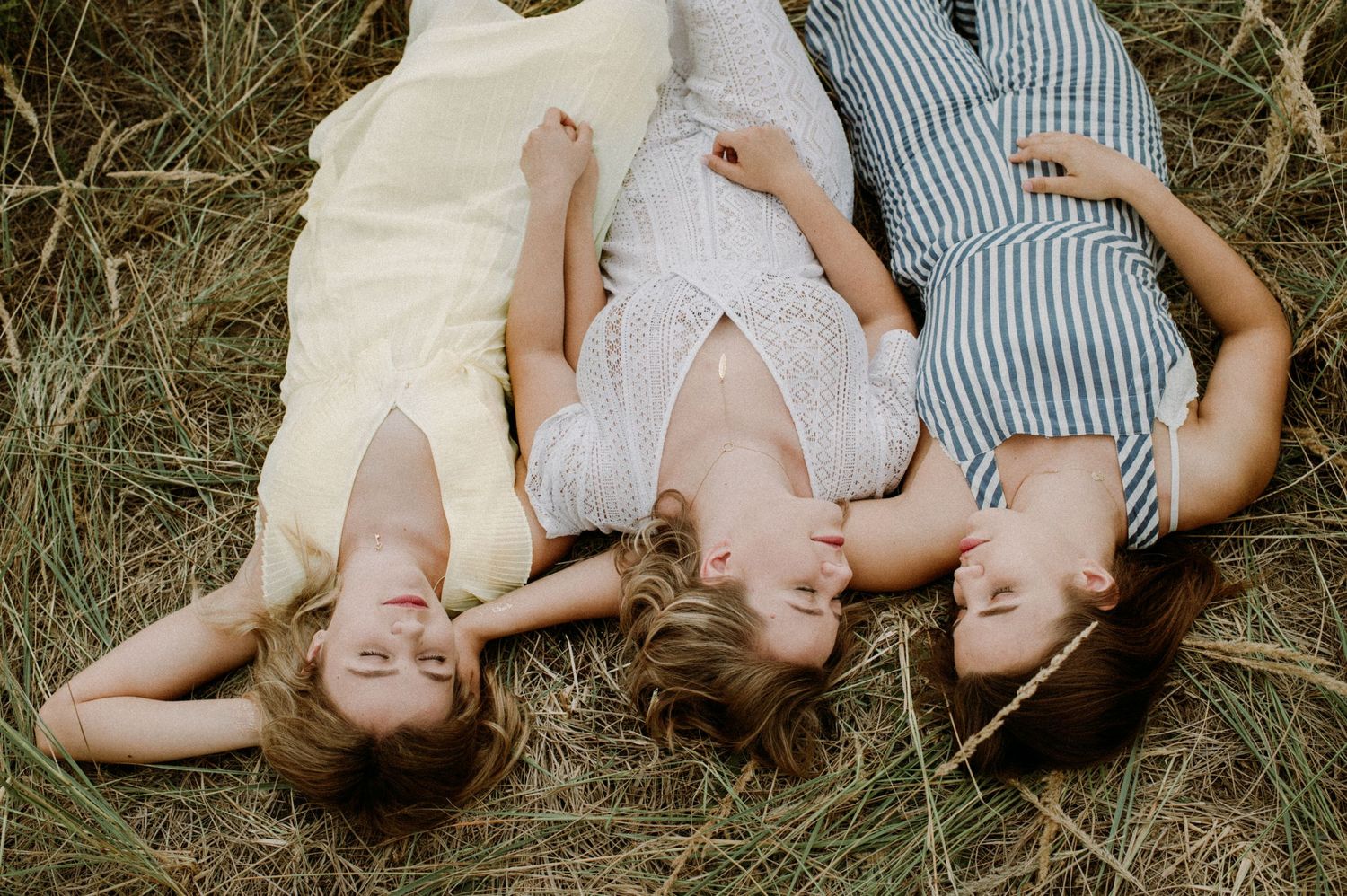 Three people lying on grass wearing light summer dresses in a candid overhead shot.