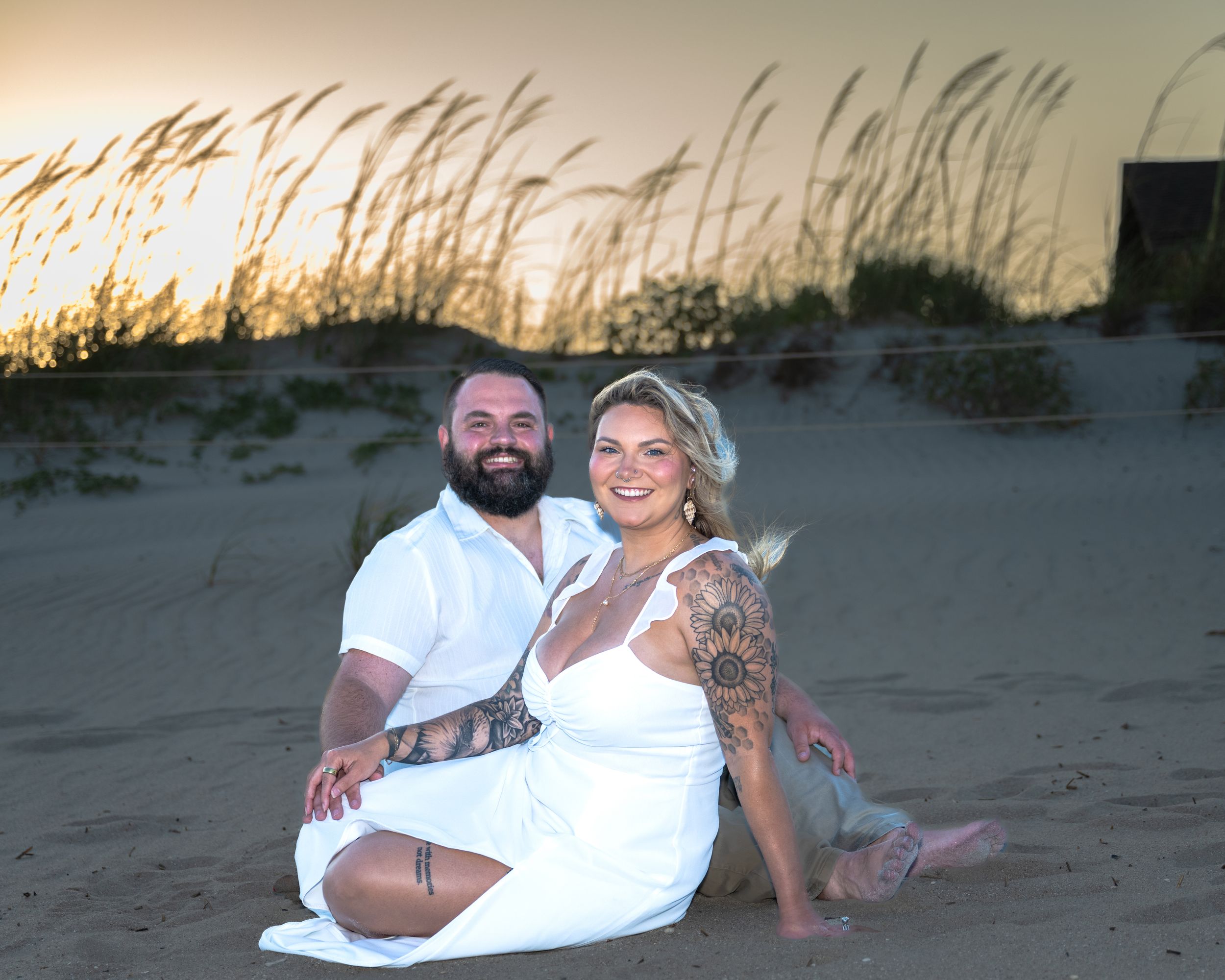 A couple sits together on a beach at sunset with sea oats swaying in the background.