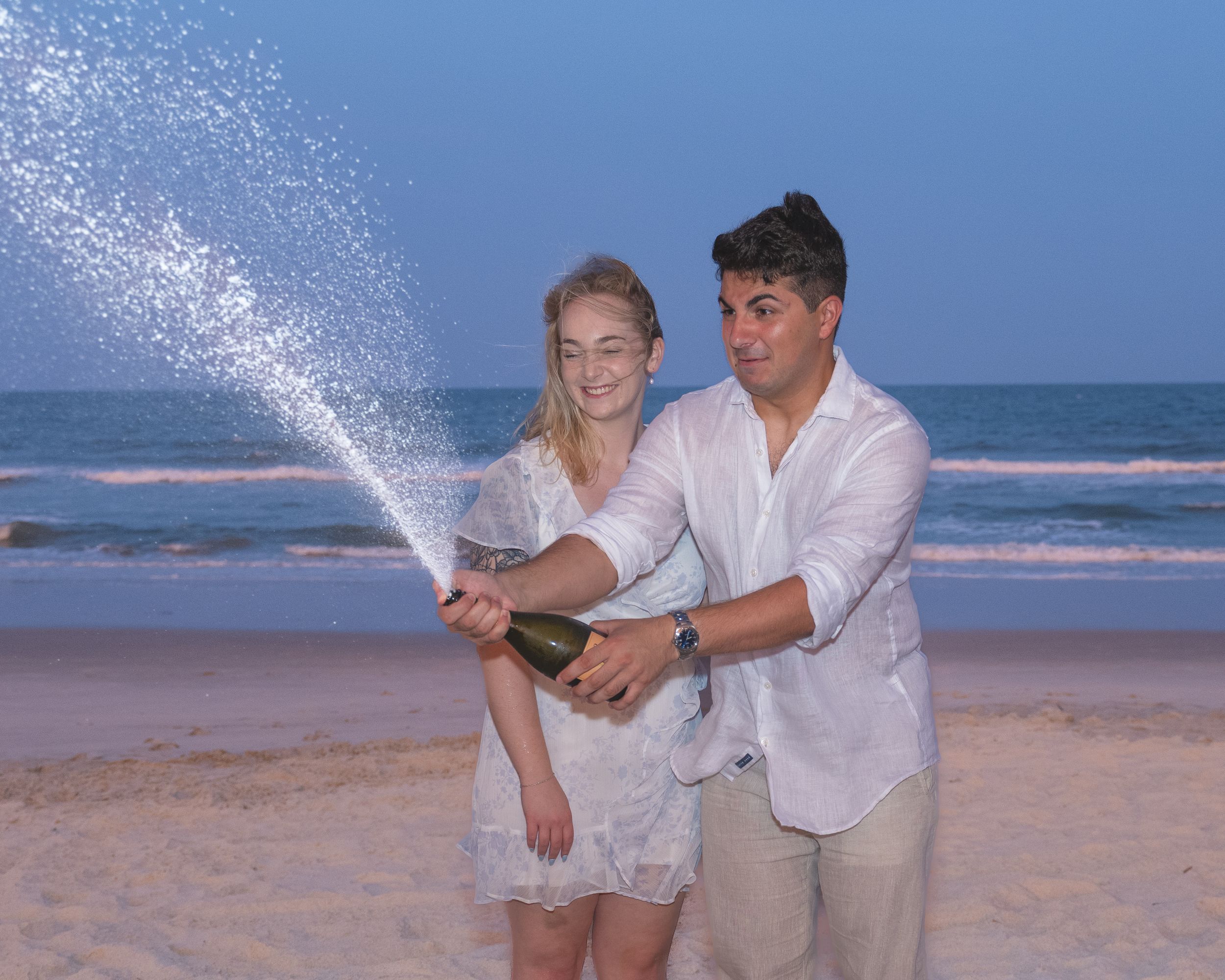 Two people in white clothing celebrating and spraying champagne on a sunny beach with ocean waves in the background.
