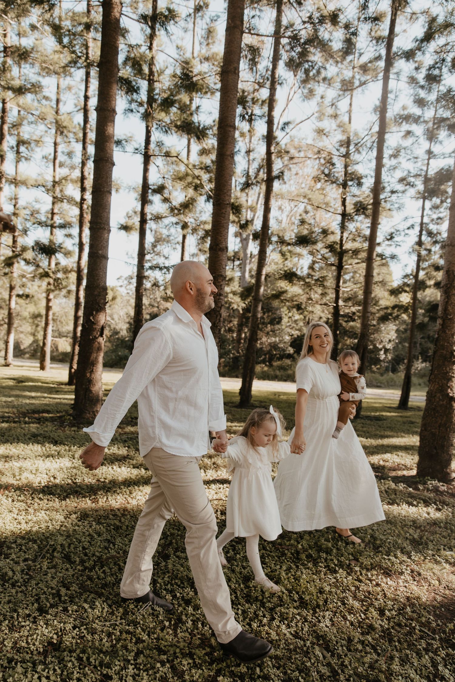 Family in white outfits playing together among tall pine trees at sunset.