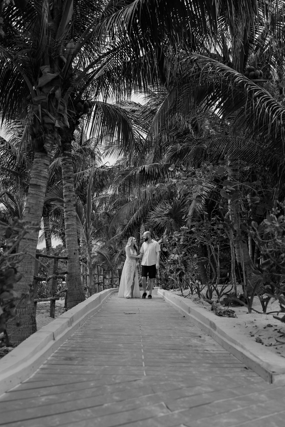 A couple walks along a wooden boardwalk under tall palm trees in a black and white tropical setting.
