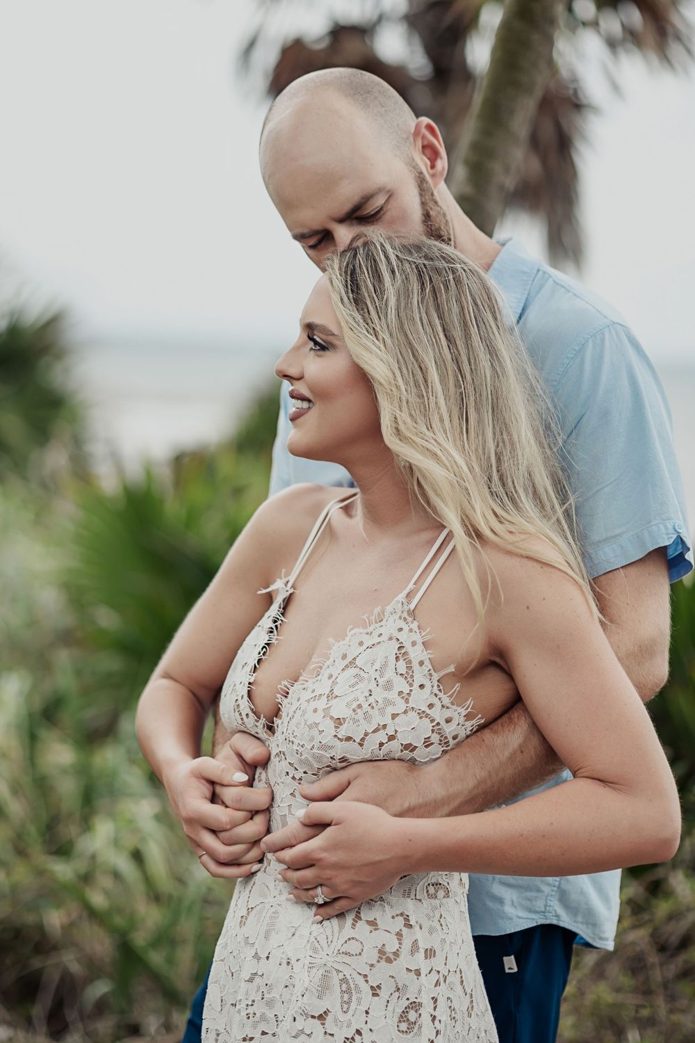 Romantic couple embraces on a beach path with palm trees, the woman wearing a delicate lace dress.