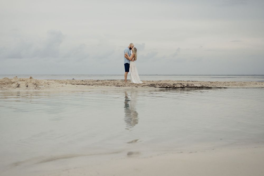 Two people stand close together on a rocky outcrop at the beach with their reflection visible in the calm water.