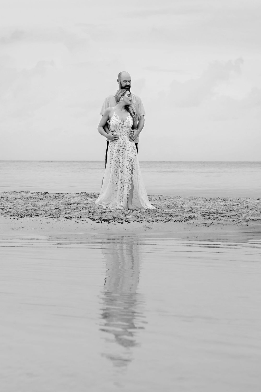Black and white photo of a couple in wedding attire embracing on a beach with their reflection in the water.