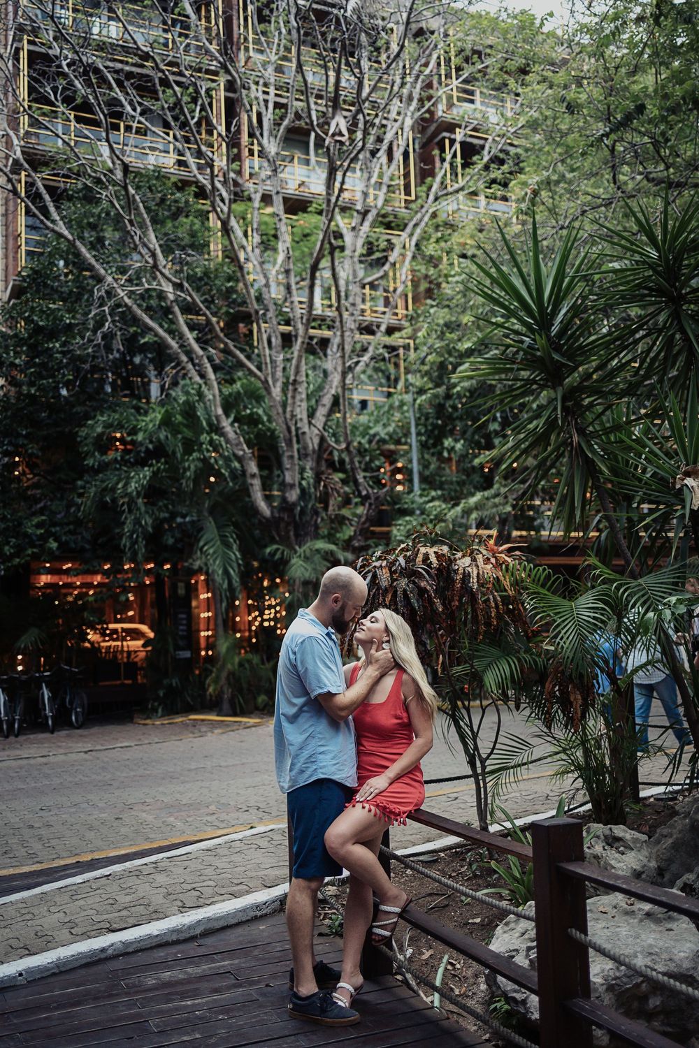 A couple share an intimate moment on a wooden deck surrounded by lush tropical plants and string lights at dusk.