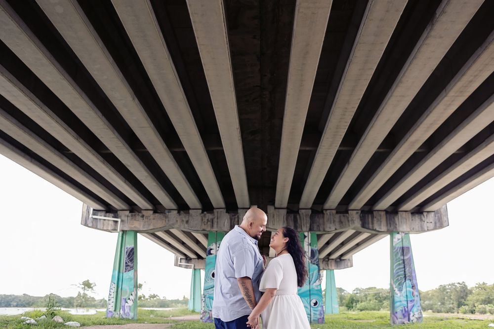 A couple stands beneath the geometric patterns of an overpass supported by turquoise columns on a sunny day.